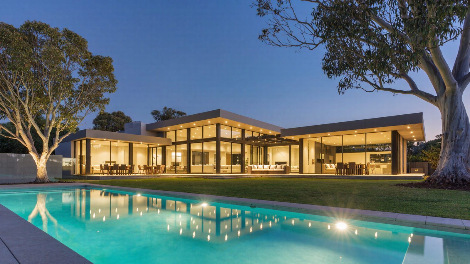 A glass pavilion home at dusk with a lit pool in the foreground
